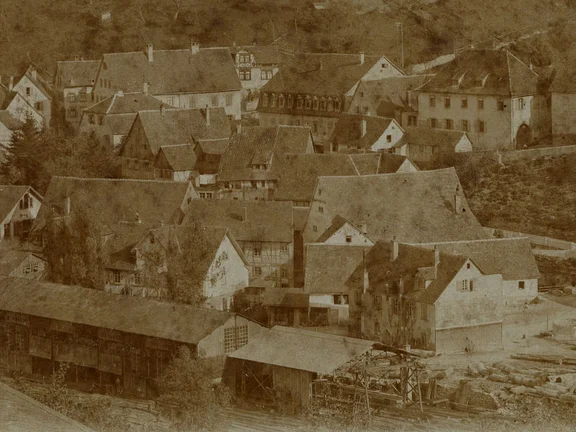 Blick aus Richtung Nordwesten auf den Bereich am Mühlkanal. Das Hauptgebäude der teilweise durch die Bauten der Sägemühle verdeckten Mahlmühle ist an der großen Dachfläche in der rechten Bildmitte erkennbar; bei dem dahinter leicht nach links versetzt sichtbaren Bauwerk handelt es sich wohl um die heutige Mühlsteige 10. Ausschnitt aus einem Vorlagenfoto aus dem Bestand des Schwäbisch Haller Postkartenverlags Seyboth, um 1900. Fotograf nicht genannt (StadtA Schwäb. Hall Seyboth F 0101)