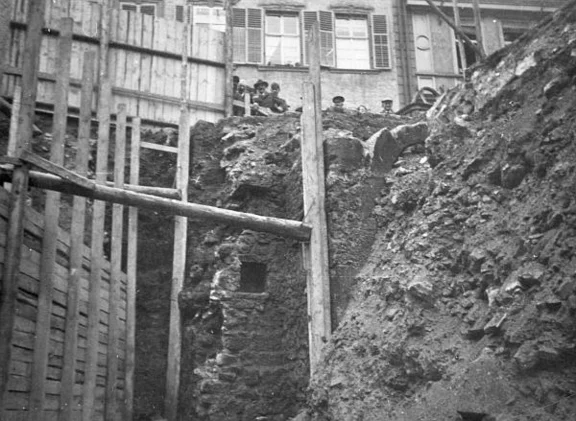 Blick aus der Baugrube auf deren südliches Ende, im Hintergrund die Häuser Haalstraße 5 (Haller Tagblatt) und Haalstraße 7. In dem wohl aus dem Mittelalter stammenden Mauerwerk gibt es eine Nische oder ein kleines Fenster, in dem rechts davon erkennbaren Mauervorsprung befindet sich ein nur teilweise freigelegtes Portal. Insbesondere zweiteres ist ein Indiz für eine deutliche Anhebung der Bodenniveaus durch Schuttablagerungen etc. – aus dem Erdgeschoss wurde auf diese Weise Kellermauerwerk. Foto: E. Schwend (StadtA Schwäb. Hall FS 41108)